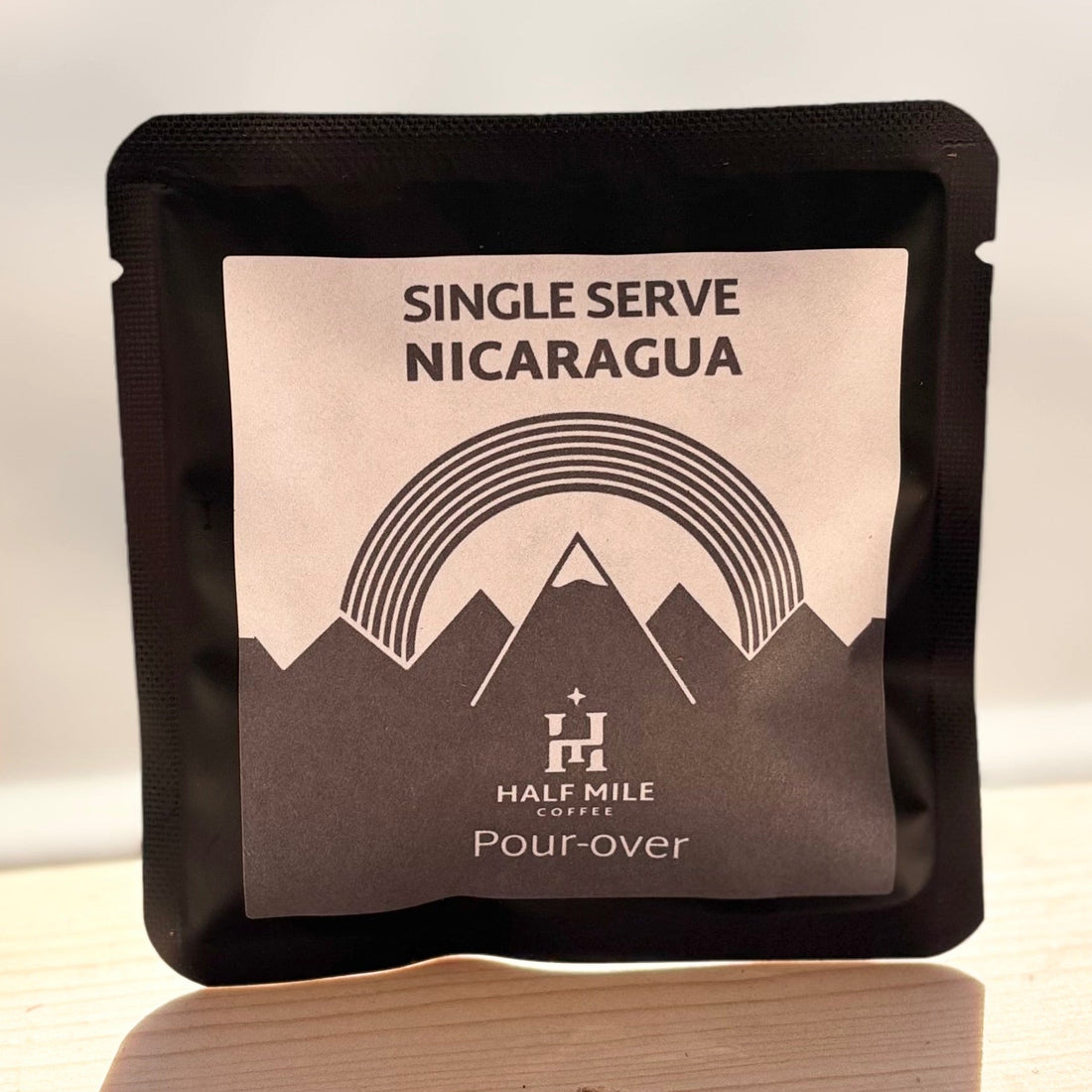 A single-serve coffee pouch from Half Mile Coffee on a light wooden surface with a white background. The pouch is black with a central label that reads 'SINGLE SERVE NICARAGUA' and features a mountain and rainbow graphic.