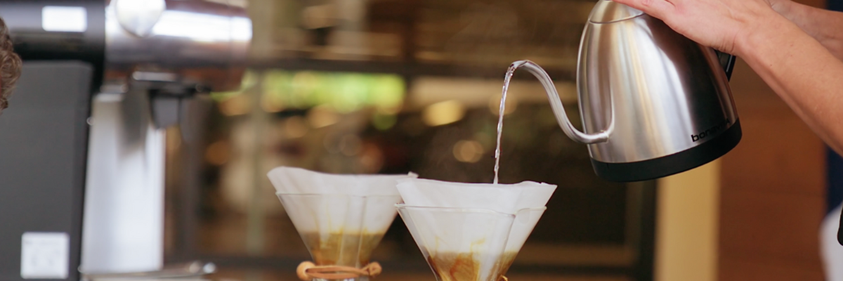 Kettle pouring hot water into two Chemex brewers with coffee and filters on a wooden counter